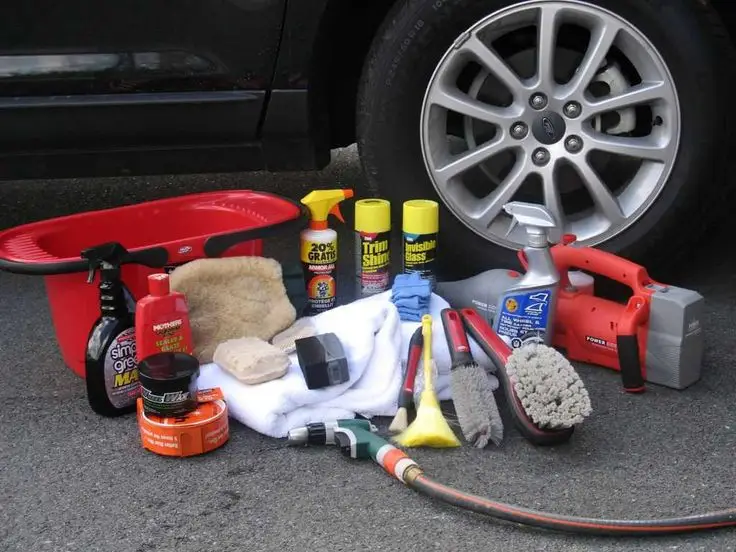 Car cleaning supplies including brushes, towels, and sprays arranged by a vehicle wheel on asphalt.