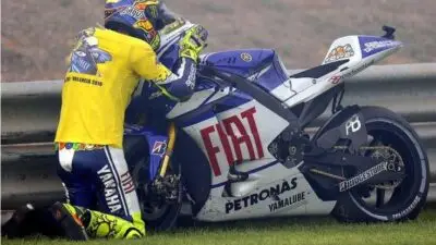 Motorcycle racer kneeling by a Fiat-branded bike on grass, wearing yellow Yamaha gear.