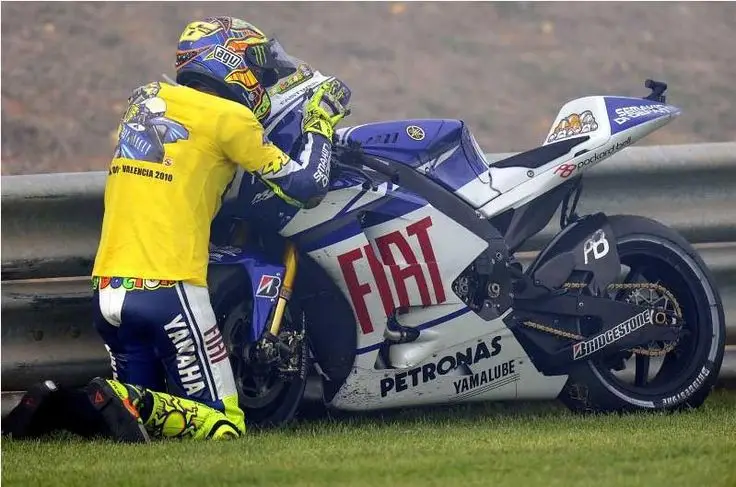 Motorcycle racer kneeling by a Fiat-branded bike on grass, wearing yellow Yamaha gear.