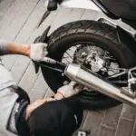 Garage mechanic working on motorcycle in China - Engine Stories Mechanic repairing motorcycle rear wheel on a brick-paved surface. Worker adjusting exhaust pipe with gloves.