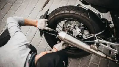 Mechanic repairing motorcycle rear wheel on a brick-paved surface. Worker adjusting exhaust pipe with gloves.