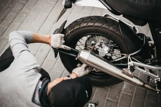 Mechanic repairing motorcycle rear wheel on a brick-paved surface. Worker adjusting exhaust pipe with gloves.