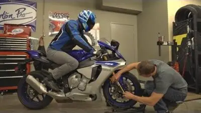 Mechanic checks tire on blue motorcycle inside a garage while rider sits on the bike, wearing helmet and gear.