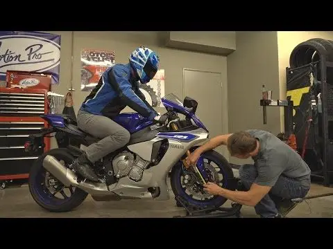 Mechanic checks tire on blue motorcycle inside a garage while rider sits on the bike, wearing helmet and gear.