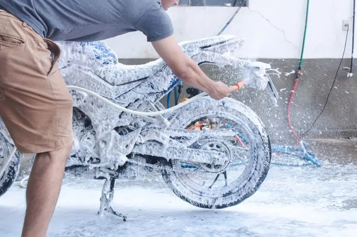 Man washing a motorcycle with soap and water at a car wash, focusing on cleaning the wheels and frame for a shiny finish.