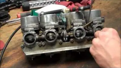 Hand tuning a motorcycle carburetor on a workbench, surrounded by tools and repair equipment.