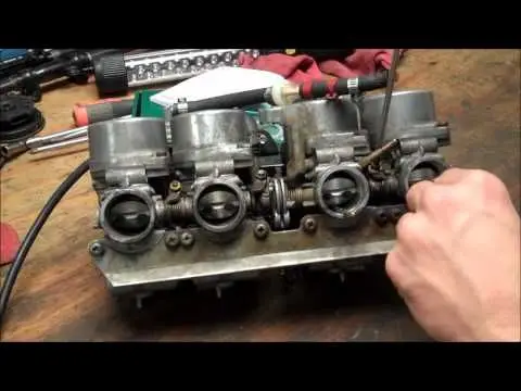 Hand tuning a motorcycle carburetor on a workbench, surrounded by tools and repair equipment.