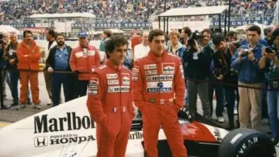 Two racecar drivers in red suits pose by a Honda Formula 1 car at a crowded race track.