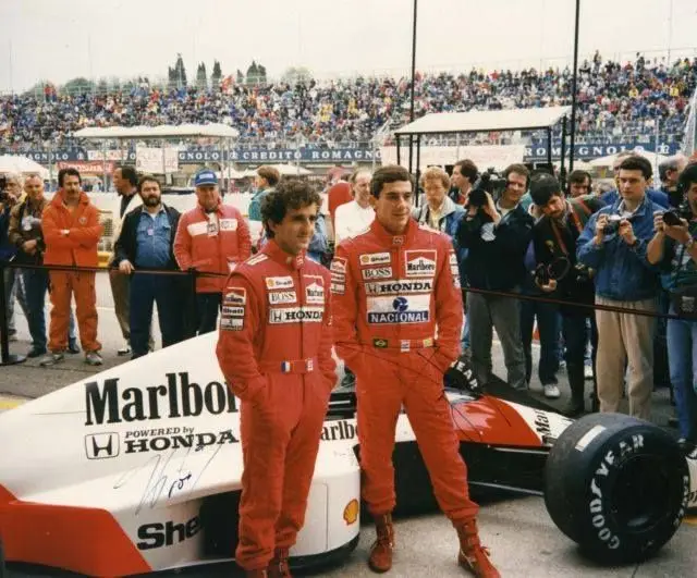 Two racecar drivers in red suits pose by a Honda Formula 1 car at a crowded race track.