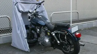 Motorcycle stored in a protective outdoor cover, parked on concrete near a chain-link fence.
