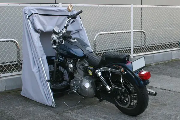 Motorcycle stored in a protective outdoor cover, parked on concrete near a chain-link fence.