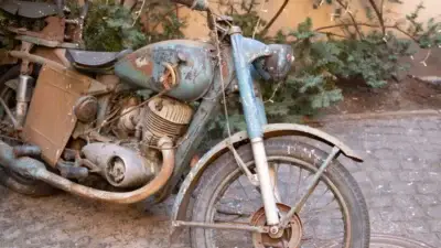 Rustic vintage motorcycle with weathered blue paint parked on cobblestone, surrounded by greenery.