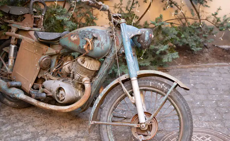 Rustic vintage motorcycle with weathered blue paint parked on cobblestone, surrounded by greenery.