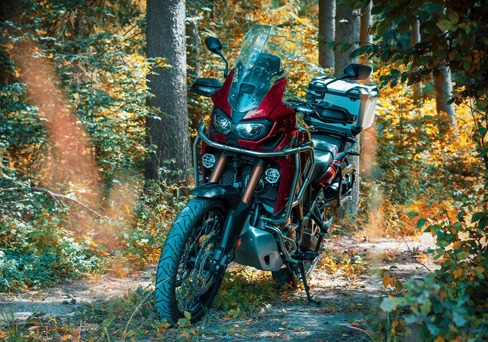 Red adventure motorcycle parked on a forest trail, surrounded by vibrant autumn foliage.