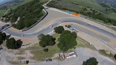 Aerial view of a winding racetrack surrounded by greenery, featuring several cars navigating a sharp turn on a sunny day.