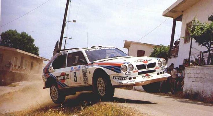 Rally car in mid-air on a dirt road, sporting Martini Racing livery. Thrilling motorsport action captured in a rural setting.