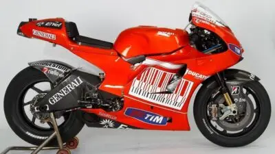 Red racing motorcycle with sponsor logos and a stand, showcasing sleek design and advanced engineering. Perfect for motorsport enthusiasts.