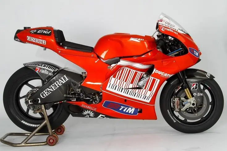 Red racing motorcycle with sponsor logos and a stand, showcasing sleek design and advanced engineering. Perfect for motorsport enthusiasts.
