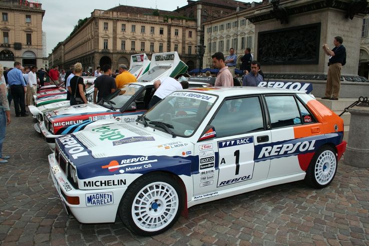 Classic rally cars displayed in a European plaza, featuring Repsol branding and car enthusiasts in the background.