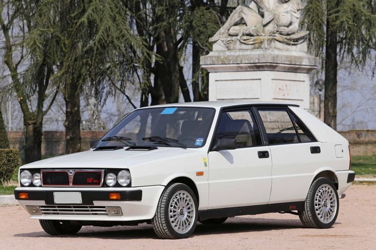 White vintage hatchback car parked in front of a statue, with trees in the background, showcasing classic design and style.