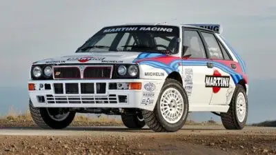 Classic rally car with Martini Racing livery on a gravel road, showcasing iconic design and performance elements.