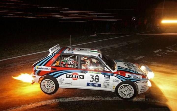 Rally car speeding at night with flames from exhaust, sharp turn, dynamic motorsport action, Martini Racing livery.