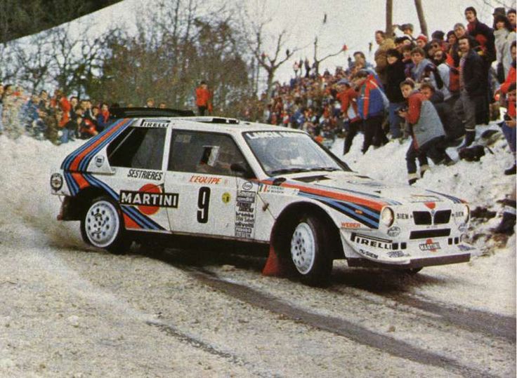 Rally car with Martini livery drifting on snow-covered track during a race, surrounded by cheering spectators.