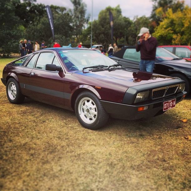 Classic maroon sports car parked at an outdoor car show with trees and spectators in the background.