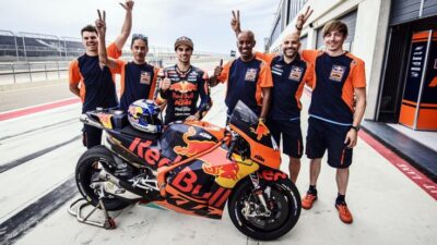 MotoGP team celebrates with rider beside Red Bull KTM bike on racetrack. Team spirit and racing success.