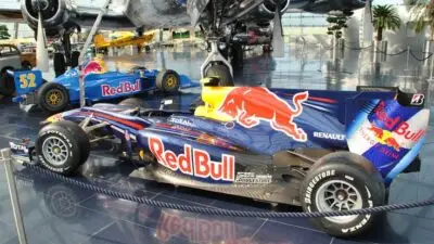 Red Bull racing cars displayed in a museum setting, showcasing sleek designs and vibrant branding under an airplane.