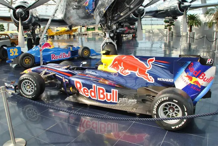 Red Bull racing cars displayed in a museum setting, showcasing sleek designs and vibrant branding under an airplane.