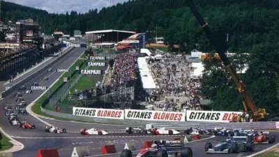 Formula 1 cars race at a historic track with stands full of spectators, featuring iconic brands and lush green surroundings.