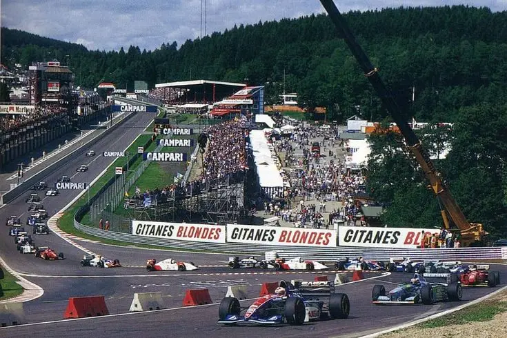 Formula 1 cars race at a historic track with stands full of spectators, featuring iconic brands and lush green surroundings.