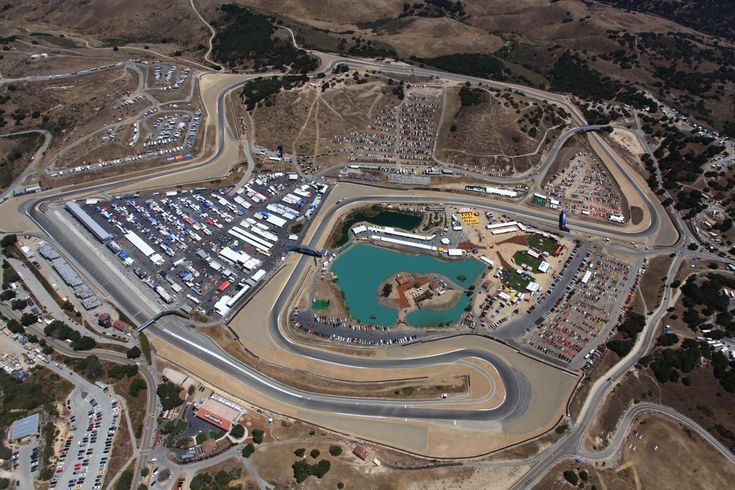 Thrilling WeatherTech Raceway Laguna Seca esports showdown - Engine Stories Aerial view of a winding race track surrounded by parked vehicles and dry terrain, with a small pond at the center.