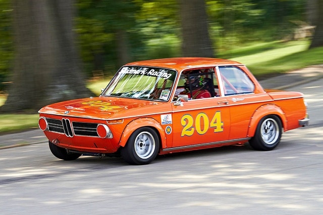 Vintage orange race car with number 204 speeding on a tree-lined road. Classic BMW model in motion at a racing event.