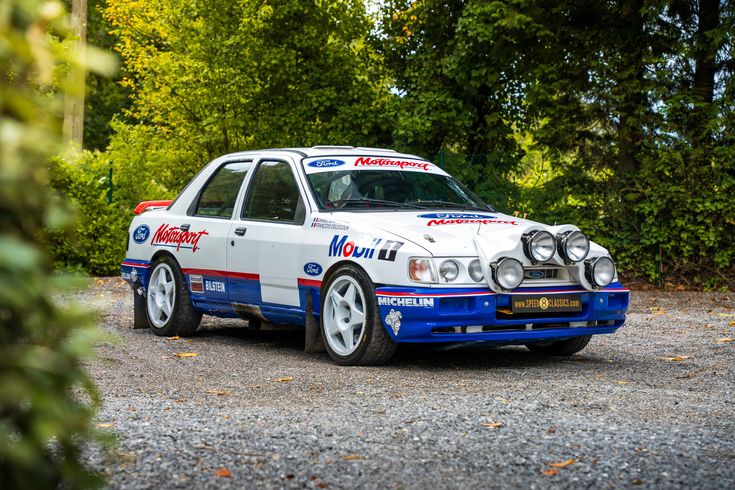 Vintage rally car with sponsor decals, parked on a gravel road surrounded by lush greenery.