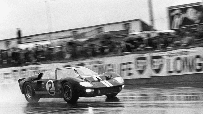 Historic black and white photo of a race car speeding on a wet track, capturing motorsport excitement and vintage racing atmosphere.