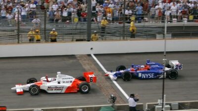 Two Indy cars racing on a track with a crowd in the background, showcasing intense competition and speed.