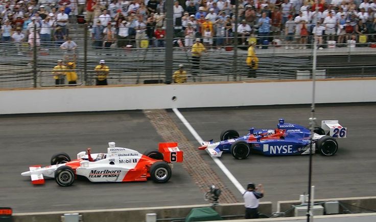Two Indy cars racing on a track with a crowd in the background, showcasing intense competition and speed.