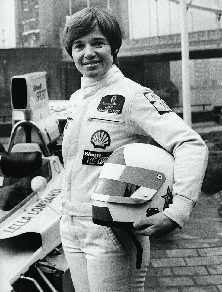 Smiling race car driver in suit holding helmet beside F1 car, showcasing passion for motorsport.