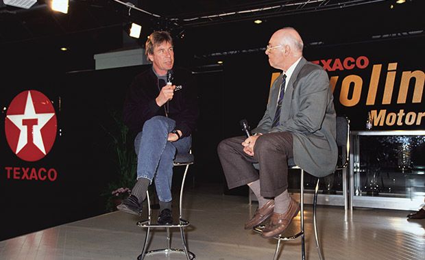 Two men seated and speaking at a Texaco event stage, with microphones in hand and Texaco logo in the background.
