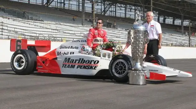 Race car driver in red suit with Marlboro Team Penske car and large trophy on a racetrack, showcasing victory celebration.