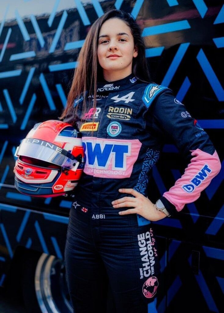 Young race car driver in BWT Alpine suit holding helmet, standing confidently.