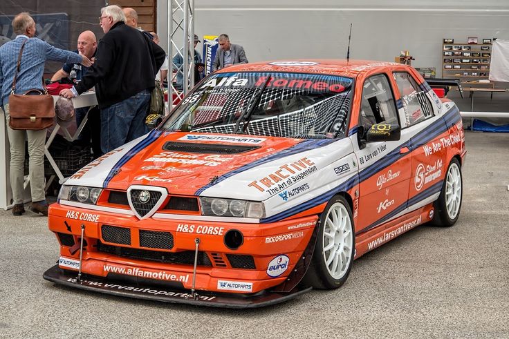 Red and blue Alfa Romeo racing car with sponsor logos at an automotive event, surrounded by people.