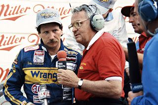 Race car driver in a Wrangler suit being interviewed by a reporter at a motorsport event, surrounded by media crew and microphones.