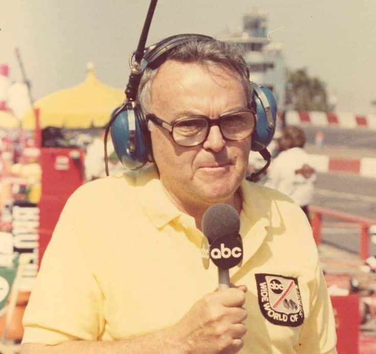 Older man with headphones holding an ABC microphone at a motorsport event. He is wearing a yellow shirt with a patch logo.