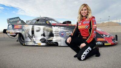 Smiling race car driver poses beside a Taylor Swift-themed dragster on a sunny day at the racetrack.