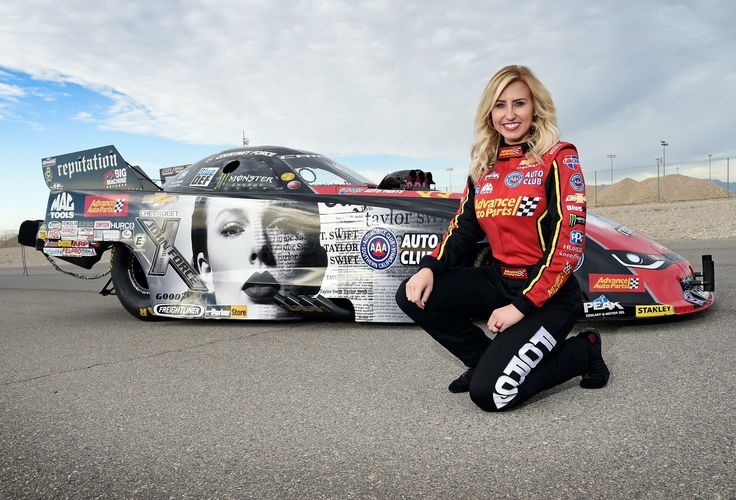 Smiling race car driver poses beside a Taylor Swift-themed dragster on a sunny day at the racetrack.
