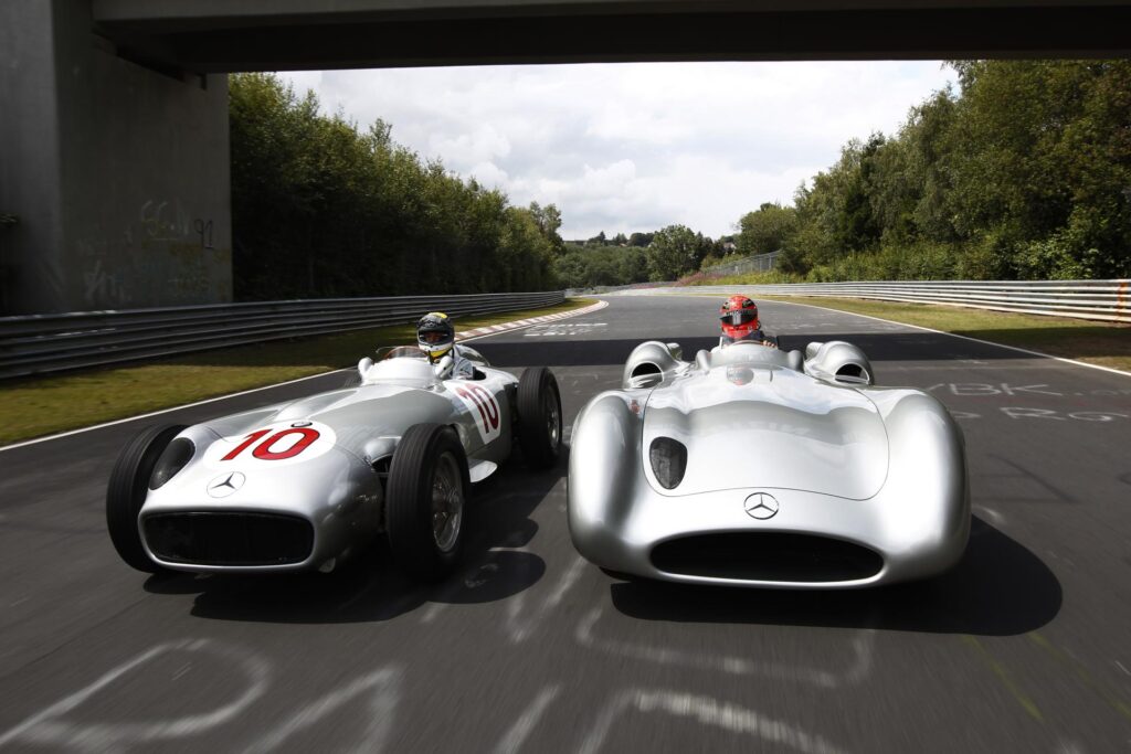 Two vintage Mercedes Silver Arrow race cars on a track, showcasing classic motorsport heritage and design under a bridge.
