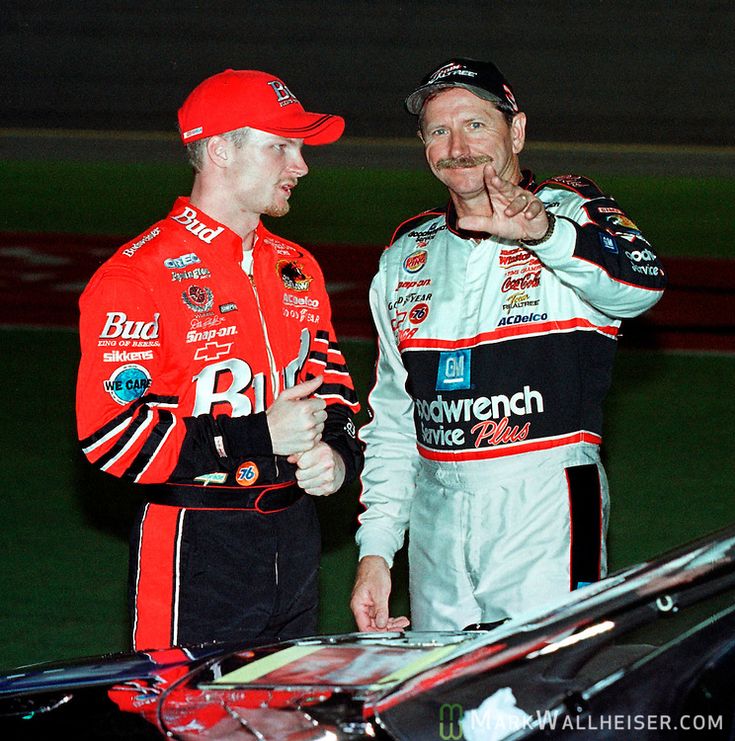 Two NASCAR drivers in racing suits engage in conversation next to a race car.